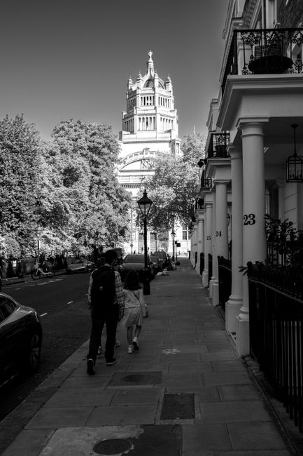 A black-and-white photograph depicts a residential street scene in West Kensington, with a couple walking along the sidewalk towards a large historic building topped with a tower and spire in the back