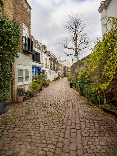 A narrow cobblestone street in West Kensington lined with residential buildings on both sides, featuring white and brick facades, some with small balconies and potted plants placed outside. There is a large, leafless tree visible against an overcast sky at the end of the street. On the right side, a tall stone wall is covered with green foliage, while the left side shows windows with white frames and various potted plants on window sills and pavement. The scene depicts an urban residential area suitable for house removals or home relocation services. Man and Van West Kensington may utilize such access points during furniture transport and packing and moving processes, where careful navigation or loading logistics might be necessary.