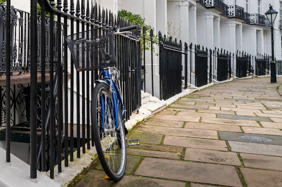 A blue bicycle with a front black mesh basket is locked to a black metal fence outside a row of white terraced houses in West Kensington. The fence features pointed finials and separates the pavement from the steps leading up to the residential entrances. The pavement is paved with large, irregular stone slabs, some showing signs of moss or dirt in the joints. Several black bollards are positioned along the edge of the pavement, and a few of the house entrances have white steps with black railings. The buildings have black wrought iron balconies and traditional street lamps attached to the walls. The scene appears to be during daylight with natural lighting, emphasizing the traditional urban setting. This image relates to house removal and moving logistics, such as the typical obstacles faced when relocating and accessing properties in the West Kensington area, which [COMPANY_NAME], such as [DOMAIN], can assist with during house removal or furniture transport services.