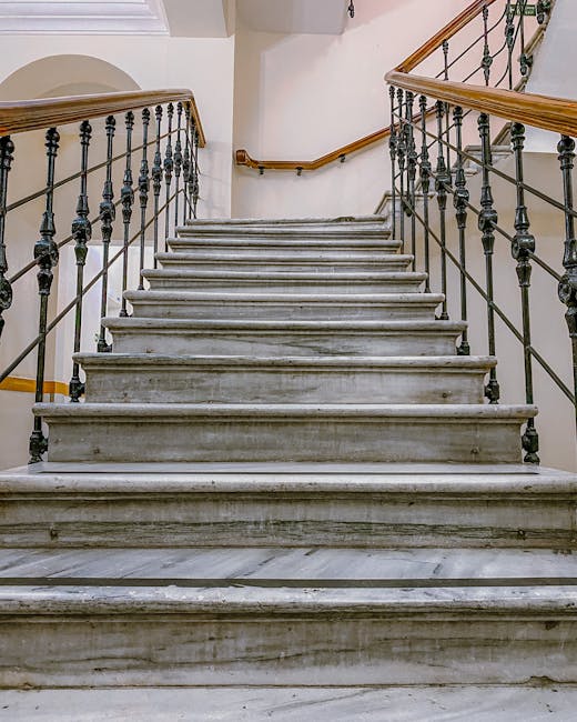 A straight staircase made of grey marble with multiple steps, situated inside a building. The staircase has black wrought iron railings with decorative elements on either side, topped with polished wo