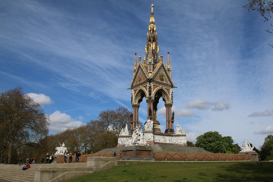 A tall, ornate monument situated on a grassy area with steps leading up to it, featuring intricate Gothic-style architecture highlighted by decorative sculptures and pointed spires, with a backdrop of a partly cloudy blue sky and surrounding trees. In the foreground, several people are visible, some walking and others standing, while a group appears to be observing or taking photographs. The structure is positioned near the pavement, indicating it is located in a park or public space. Although the primary focus is on the monument, the image context suggests a setting associated with city landmarks or historical sites. No furniture, boxes, or moving equipment are visible in this image, but the scene's open outdoor environment aligns with the idea of a location where home relocation or sightseeing may take place, and [COMPANY_NAME] may occasionally be involved in logistics or transportation related to such areas.