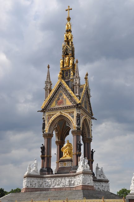 A detailed view of the ornate national monument situated outdoors against a cloudy sky, featuring a Gothic-style spire adorned with gold accents, intricate mosaics, and statues. The structure rests on a circular base decorated with sculptural reliefs and classical figures, with an arched canopy supported by columns framing a golden statue at the center. Surrounding the base are additional white sculptures and decorative elements. Inside a property, the scene depicts a home relocation process with nearby cardboard boxes, protective wrapping on furniture, and a man using a trolley or straps to assist with loading items into a large van parked outside. The environment suggests an organized packing and moving operation, facilitated by professional removal services like Man and Van West Kensington, supporting efficient furniture transport and packing during a house move in the West Kensington area.