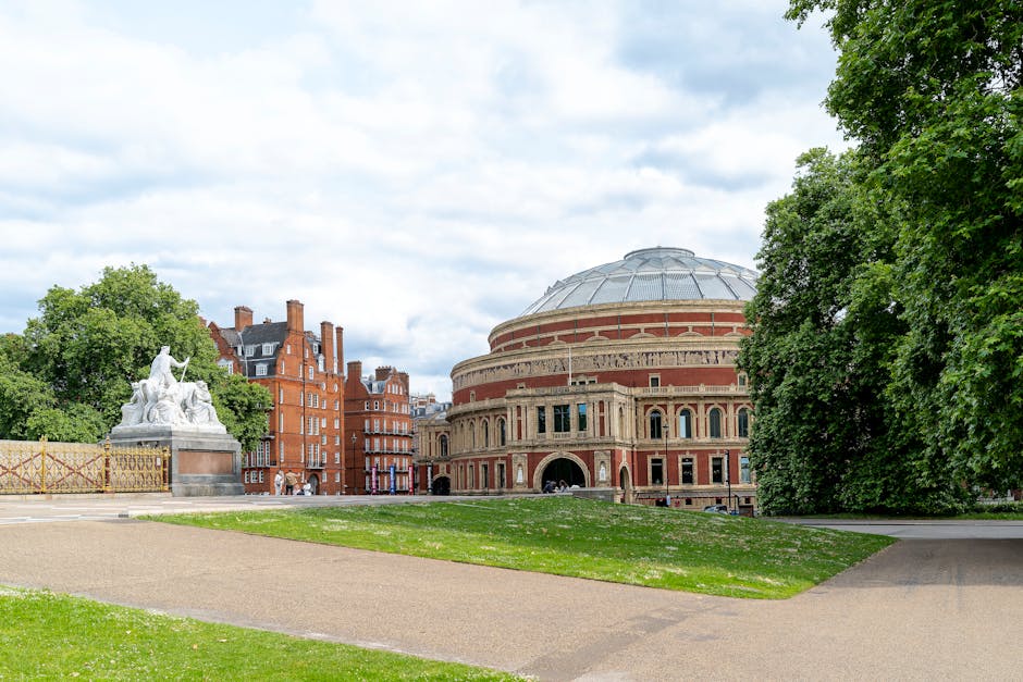 A wide outdoor view of the Royal Albert Hall in the Royal Borough of Kensington and Chelsea, showing its distinctive round, domed brick architecture with arched windows and decorative detailing. In the foreground, a paved walkway curves around a grassy area with some scattered white flowers. To the left, a white marble statue depicting a seated figure with a raised arm is situated on a stone pedestal, partially obscured by a wooden barrier with yellow tape. Tall, leafy green trees border the scene on the right and behind the statue, providing a natural frame and shade. The sky above is partly cloudy with patches of blue, and the surrounding area is calm with no visible traffic or crowds. For house removals and furniture transport, [COMPANY_NAME] often manages the loading process here, ensuring careful packing and handling of household items during home relocation projects in the vicinity.