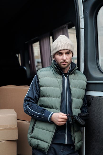 A man stands inside the open rear of a moving van, equipped with cardboard boxes of various sizes, some wrapped in plastic and others in plain brown cardboard, ready for furniture transport during a home relocation. He is wearing a light-colored knit beanie, a padded green and navy blue jacket, and black gloves, suggesting cold weather. The van's interior has a dark fabric ceiling and a sliding side door visible in the background. The boxes are stacked neatly, and the man appears to be preparing for or in the process of loading or unloading items, supporting services related to packing and moving with Man and Van West Kensington, as indicated by the webpage title and related context.