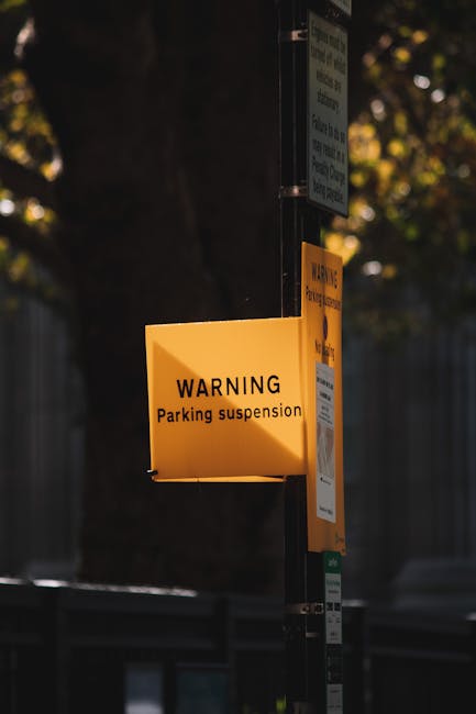 A yellow rectangular warning sign attached to a metal pole indicating 'Parking suspension' in black text, mounted next to other smaller signs on a street in West Kensington. The background includes a tree with leaves illuminated by natural daylight and a part of a fence, suggesting an urban residential area. The sign's placement is relevant for house removals and moving logistics, and Man and Van West Kensington may operate in this area for local moves, emphasizing the importance of parking permits and suspension notices for efficient furniture transport and home relocation activities.