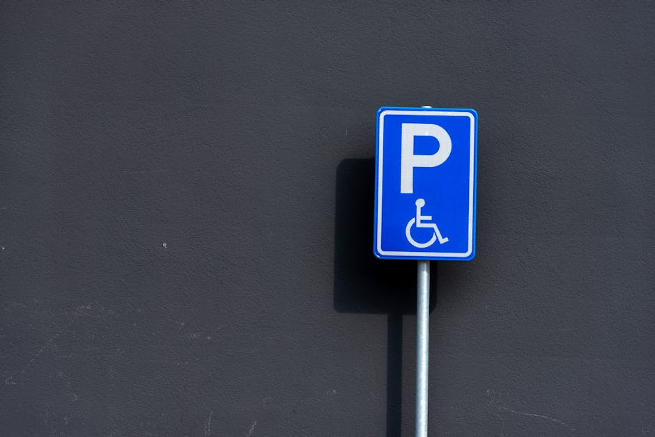 A blue parking permit sign with a white letter 'P' and a wheelchair symbol is mounted on a metal post against a dark grey exterior wall. The sign indicates accessible parking, typically designated for disabled individuals, and is situated outdoors, likely on a pavement or driveway adjacent to a building. The shadow cast by the sign suggests bright lighting, and the plain wall background emphasizes the sign’s visibility. For house removals and furniture transport, such parking permits are relevant for ensuring legal parking space during a home relocation. Man and Van West Kensington may provide guidance on parking permits for local moves, as illustrated by the availability of accessible parking options in the area.