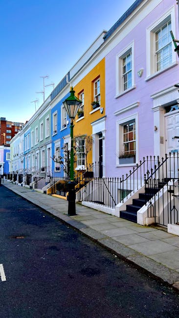 A row of historic red-brick residential buildings located on a quiet street during daytime, featuring large sash windows, decorative white trim, and small front gardens with iron fences. The buildings