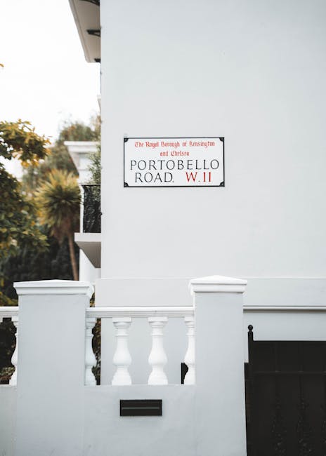 A white residential building with a small front entrance area featuring a low wall and decorative balusters, situated on Portobello Road in West Kensington. The wall surrounds a porch or small yard, with a black mailbox attached to the low wall. A rectangular street sign mounted on the building's exterior wall indicates the location as Portobello Road, W.11, positioned near the intersection of the street and the property. The setting is outdoors during daylight, with some greenery including trees visible on the left side of the image. The scene depicts a typical house exterior suitable for a home relocation or moving company to reference during packing and furniture transport activities, with clean and well-maintained features consistent with residential move logistics.