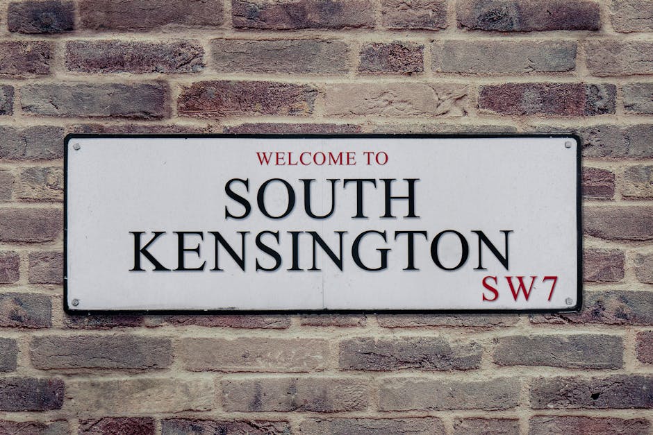 A white residential building with a small front entrance area featuring a low wall and decorative balusters, situated on Portobello Road in West Kensington. The wall surrounds a porch or small yard, with a black mailbox attached to the low wall. A rectangular street sign mounted on the building's exterior wall indicates the location as Portobello Road, W.11, positioned near the intersection of the street and the property. The setting is outdoors during daylight, with some greenery including trees visible on the left side of the image. The scene depicts a typical house exterior suitable for a home relocation or moving company to reference during packing and furniture transport activities, with clean and well-maintained features consistent with residential move logistics.