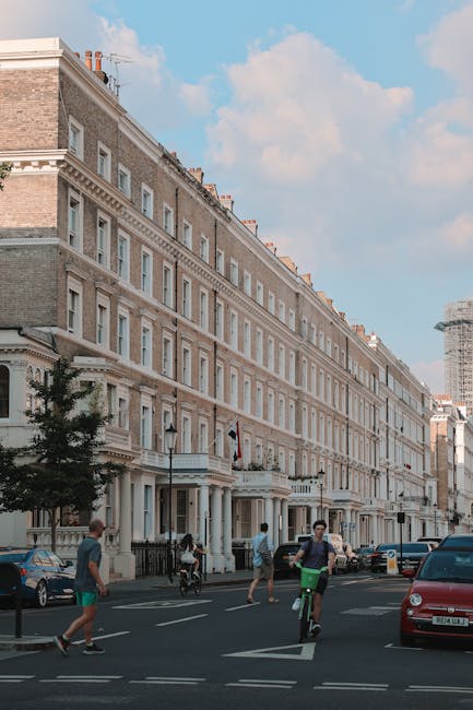 A street scene in West Kensington featuring a row of multi-storey Victorian-style terraced buildings constructed from light-colored brick and stone, with decorative cornices and sash windows. The buildings are positioned along a paved pavement with street lamps and small trees lining the sidewalk. In the foreground, several people are visible: two individuals riding bicycles, one person walking, and another walking near parked cars, including a red vehicle. The sky is partly cloudy with patches of blue. The image captures the typical residential architecture and urban environment associated with house removals and moving services in West Kensington, with Man and Van West Kensington visible as a professional local moving company supporting home relocation and furniture transport in the area.