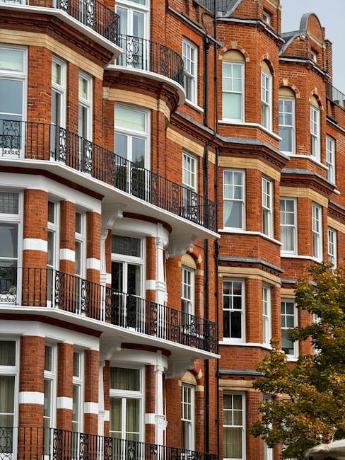 A multi-storey residential building constructed with red brick and white stone detailing, featuring large sash windows and decorative wrought iron balconies. The lower floors have curved balconies supported by white columns, with black metal railings running along the edges. The building's architectural style suggests a Victorian or Edwardian era design, situated on a tree-lined street with partial foliage visible on the lower right side. The exterior appears well-maintained, and natural lighting highlights the textured brickwork and window frames. This setting is relevant to house removals and furniture transport, capturing the type of property involved in relocation services like those offered by Man and Van West Kensington, especially during home relocation or packing and moving processes in West Kensington.