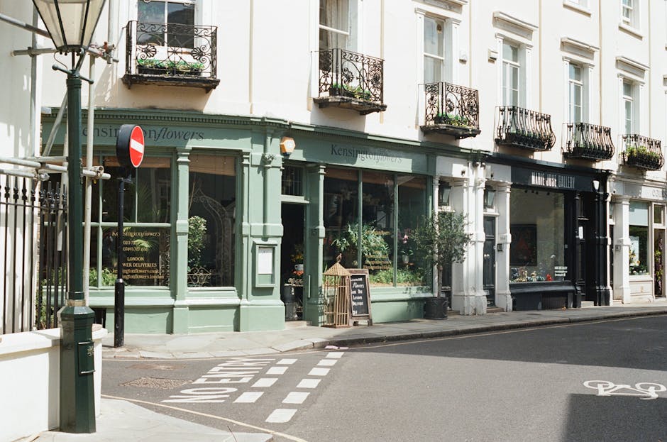 A street scene in West Kensington featuring a row of multi-storey Victorian-style terraced buildings constructed from light-colored brick and stone, with decorative cornices and sash windows. The buildings are positioned along a paved pavement with street lamps and small trees lining the sidewalk. In the foreground, several people are visible: two individuals riding bicycles, one person walking, and another walking near parked cars, including a red vehicle. The sky is partly cloudy with patches of blue. The image captures the typical residential architecture and urban environment associated with house removals and moving services in West Kensington, with Man and Van West Kensington visible as a professional local moving company supporting home relocation and furniture transport in the area.